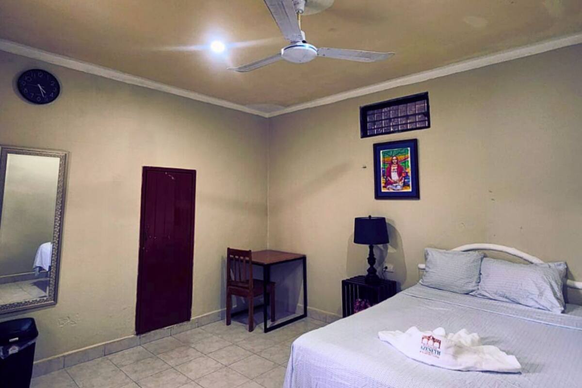 Ein Hotelzimmer mit Bett, Deckenventilator, Schreibtisch, Stuhl, Nachttisch, Lampe, einer Uhr und einem gerahmten Bild an der Wand.