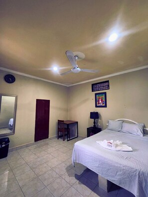 Une chambre d’hôtel comprenant un lit, un ventilateur de plafond, un bureau, une chaise, une table de chevet, une lampe, une horloge et un tableau encadré au mur.