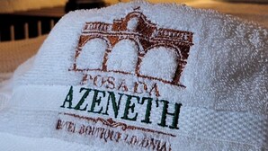 A neatly folded towel with embroidered text and a logo, placed on a bed with a visible headboard and a lamp in the background. - Posada Azeneth (Oxkutzcab)