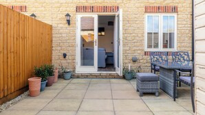 A patio with a wooden fence, potted plants, a wicker table with chairs, and a sliding glass door leading to an interior room. - Lovely 2 BD Home in Moreton-in-Marsh (Moreton-in-Marsh)