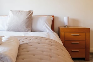 A neatly made bed with a white duvet and pillows, a wooden nightstand with drawers, and a bedside lamp. - Lovely 2 BD Home in Moreton-in-Marsh (Moreton-in-Marsh)