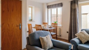 A room with a wooden door, a dining table with chairs, a sofa, and a window with curtains. - Lovely 2 BD Home in Moreton-in-Marsh (Moreton-in-Marsh)