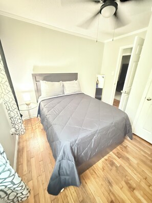Un dormitorio con cama, suelo de madera, ventilador de techo y una puerta que da a otra habitación.
