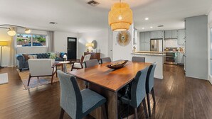 Un salon spacieux avec une grande table à manger en bois, des chaises à revêtement gris, un luminaire suspendu en rotin et une vue sur l’extérieur.