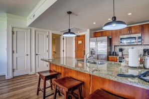 Une cuisine dotée de plans de travail en granit, d’armoires en bois, d’appareils électroménagers en acier inoxydable et de luminaires suspendus.