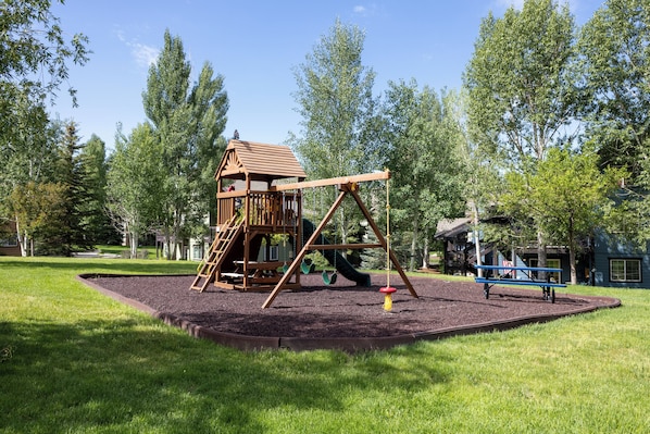 A playground with a wooden playhouse, swings, and a slide. - Quail Run 3315 - Renovated Condo, Hot Tub, Garage, W/D (Steamboat Springs)