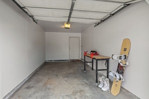 A garage with a concrete floor, a table with a snowboard and shoes, and a door in the background. - Quail Run 3315 - Renovated Condo, Hot Tub, Garage, W/D (Steamboat Springs)