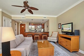 A living room with a sofa, coffee table, TV, and a kitchen area with a bar. - Honua Kai | Ocean View 3 BR, Sleeps 10 | Car Incl. w/ 6+ Nights | HKH ML-2723 by KBM (Lahaina)