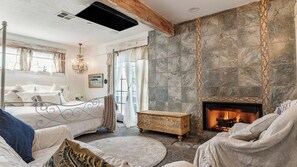 Ein Schlafzimmer mit Kamin aus Stein, einem Bett mit weißer Bettwäsche, einer Chandelierleuchtre und Blick ins Freie.