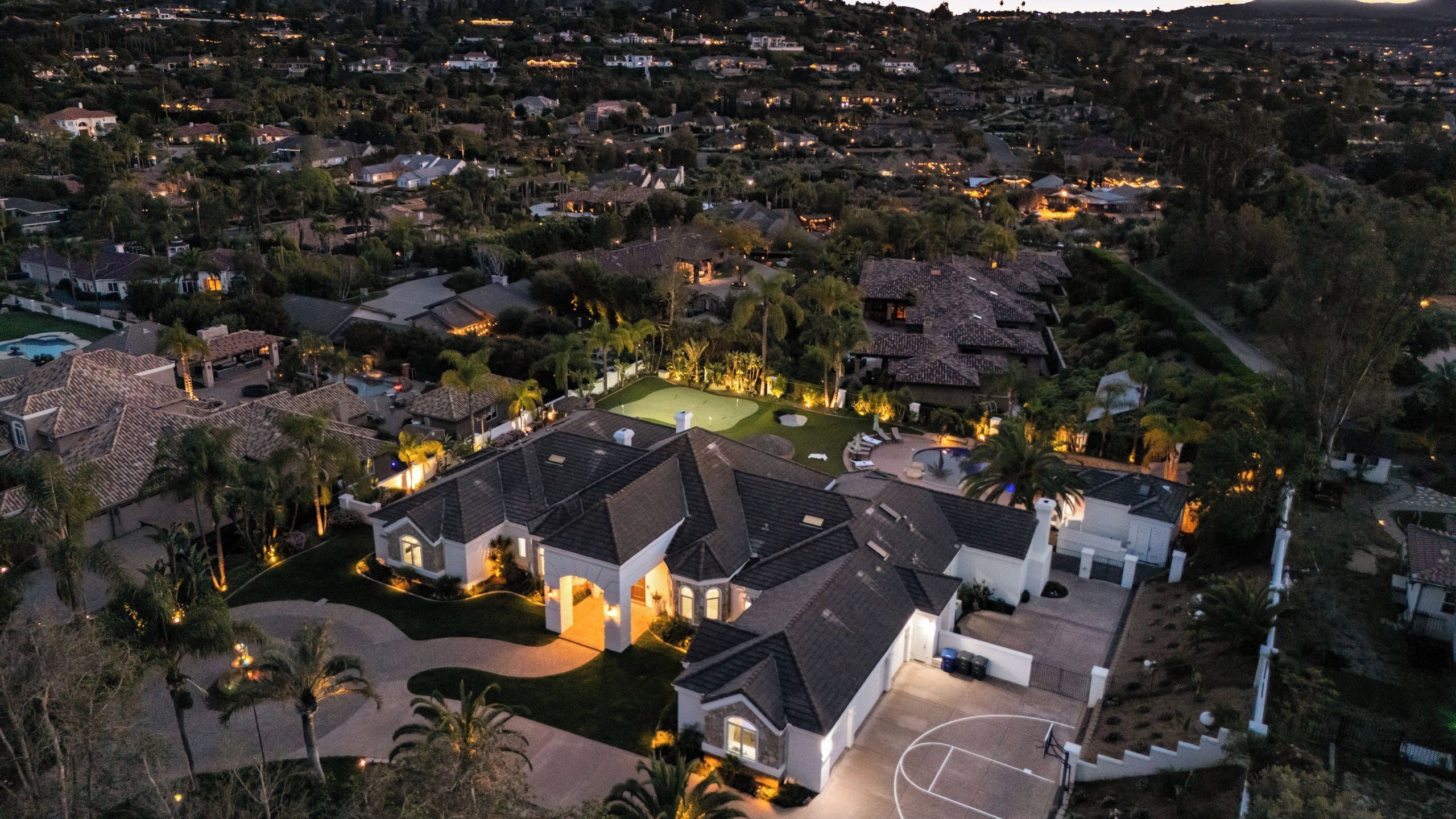 An aerial view of a residential area with houses, a swimming pool, and a well-lit tennis court.
