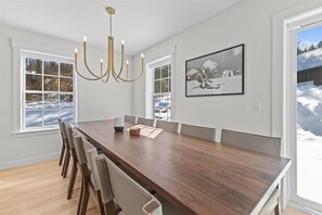 A dining area with a wooden table, chairs, a chandelier, and large windows with a snowy view. - Modern Stowe Retreat: New Build near Skiing&Town (Stowe)