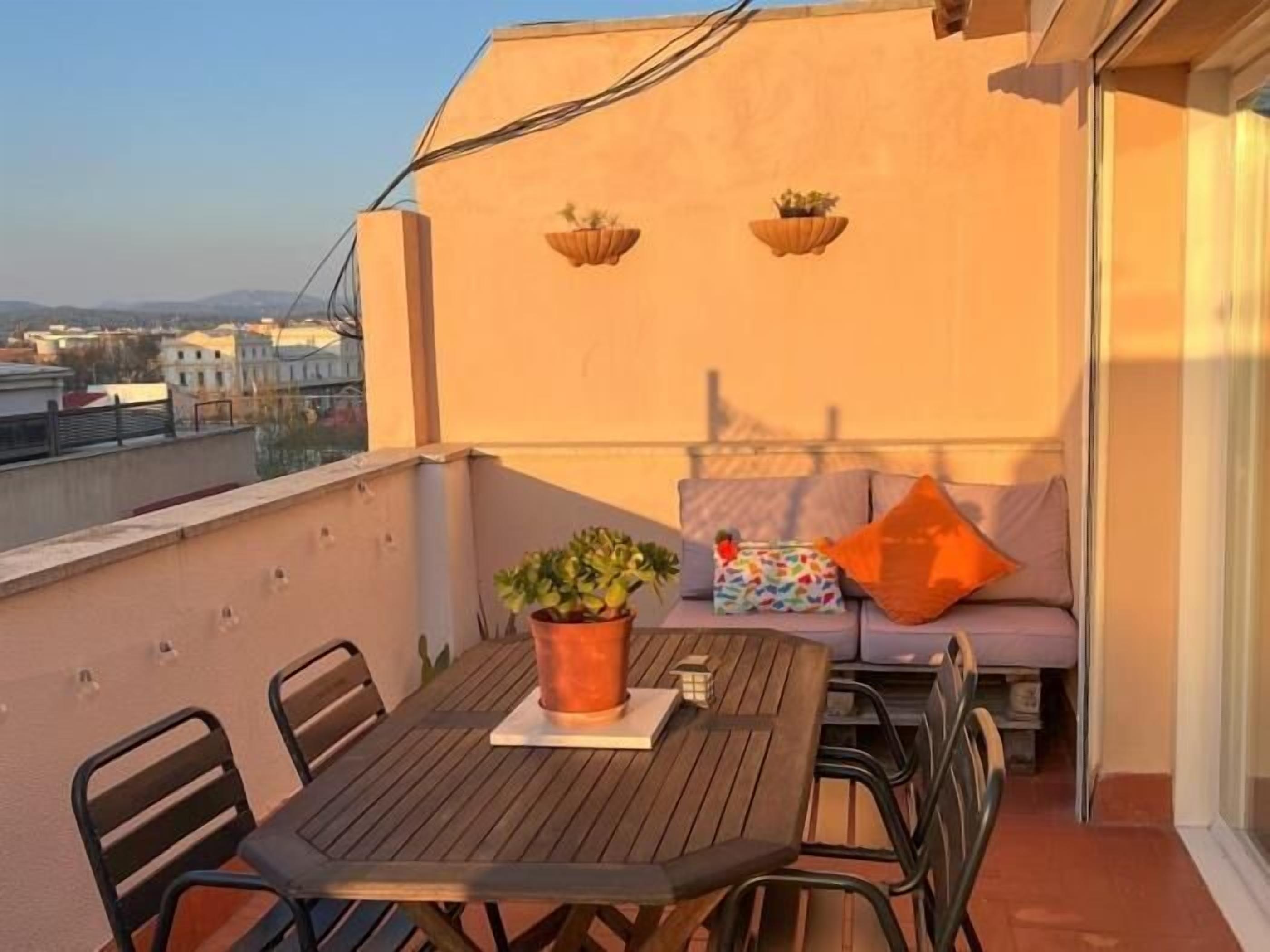 Un balcon avec une table et des chaises en bois, une plante en pot, un coin salon avec des coussins, et des jardinières suspendues.