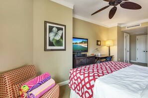 Ein Hotelzimmer mit einem Bett, einem Schreibtisch, einem Fernseher, einem Deckenventilator und einem gerahmten Bild an der Wand.