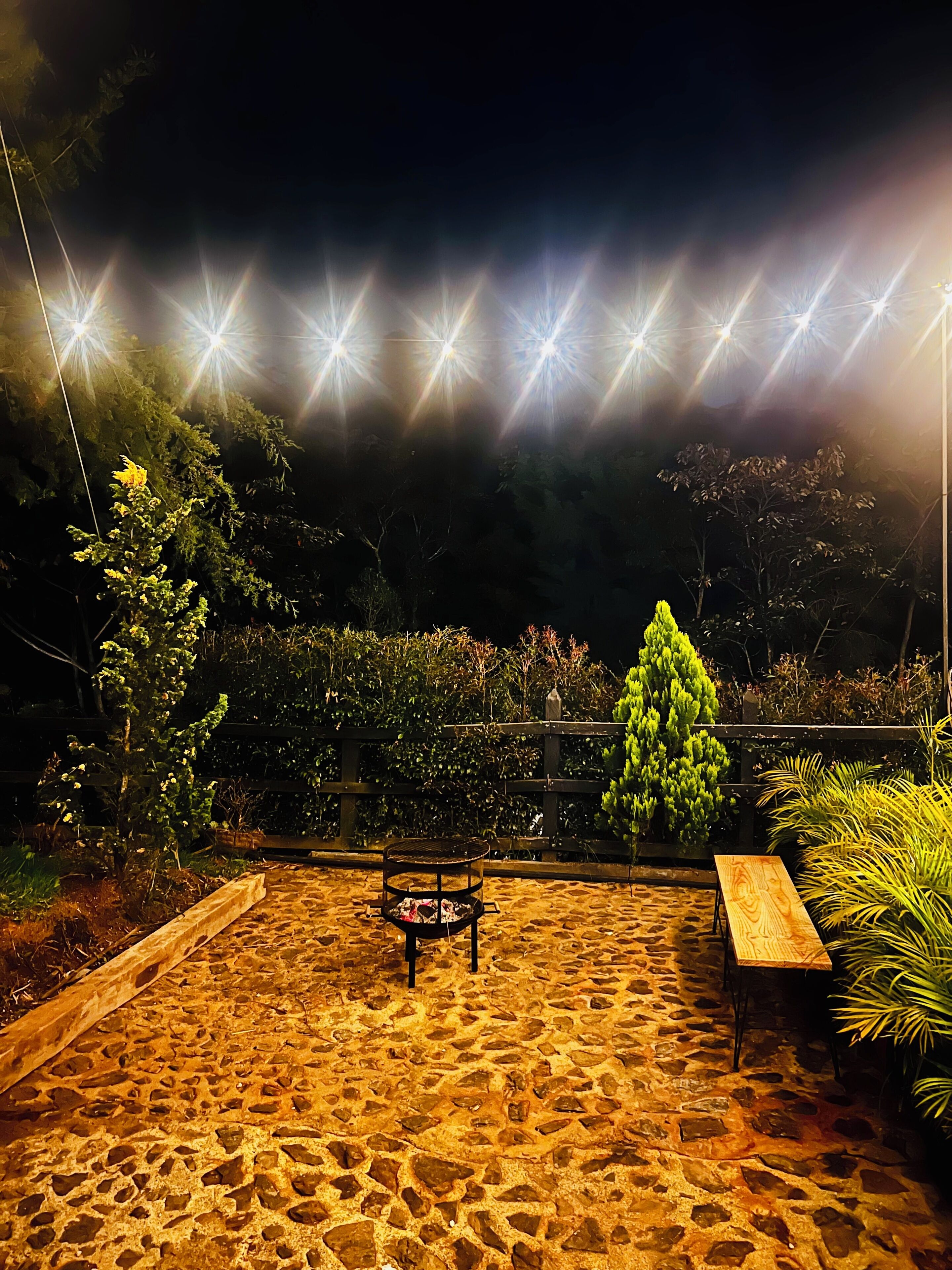 A stone-paved outdoor area with a bench and a fire pit, surrounded by greenery and a wooden fence.