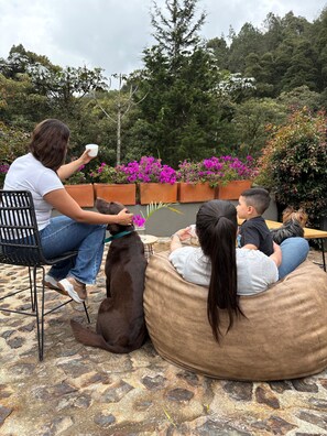 A woman sitting on a chair, a child on a bean bag, and a dog on the ground, with a garden and potted plants in the background. - Casa de campo cerca al aeropuerto MDE (Rionegro)