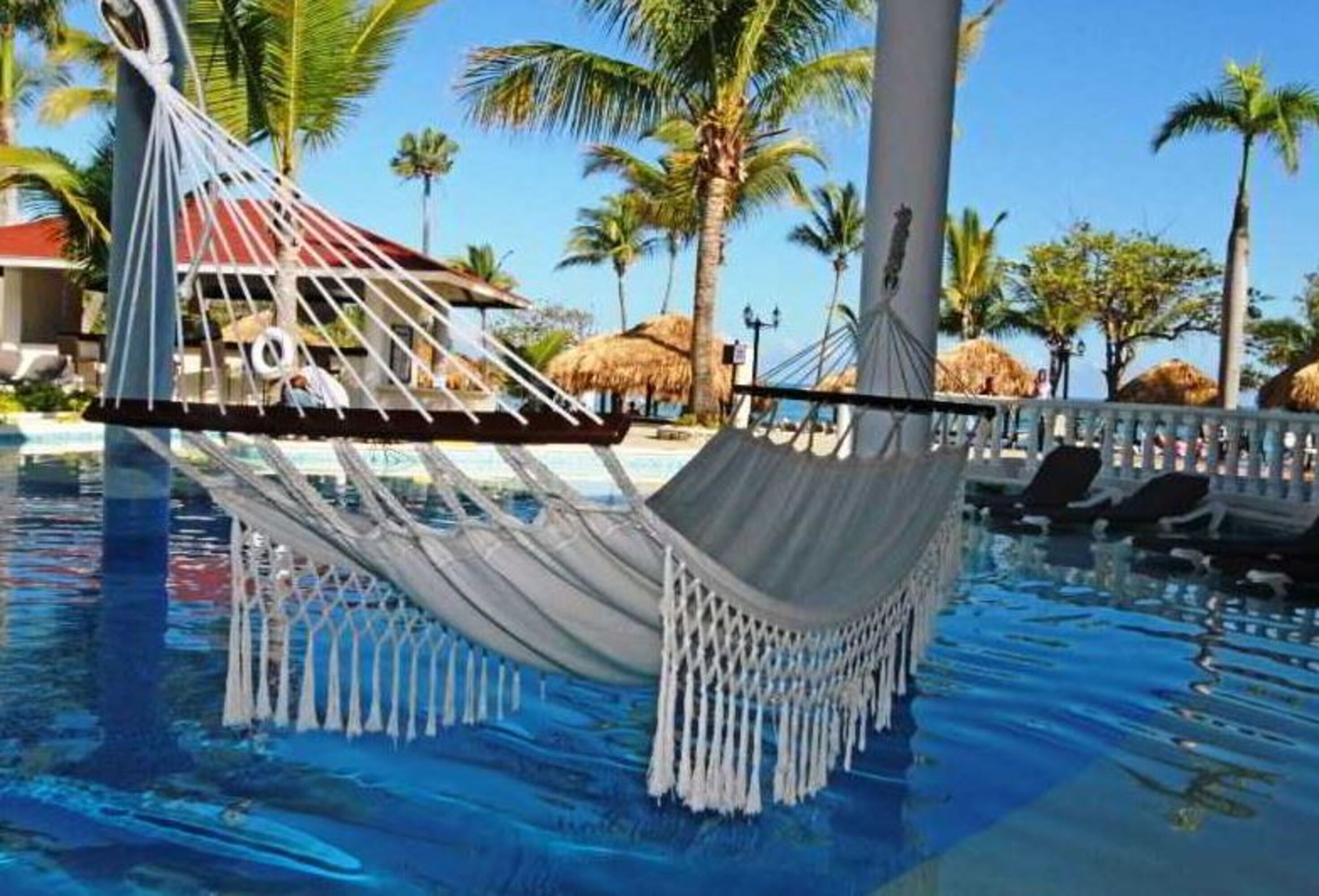 A hammock over a pool with palm trees and a thatched-roof structure in the background.