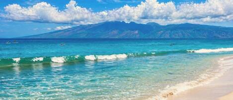 A beach with clear turquoise water, a sandy shore, and a mountain range in the background.