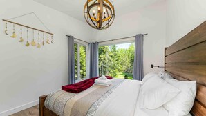 Ein Schlafzimmer mit Bett, einer Hängeleuchte, Blick auf Bäume durch das Fenster und einem Holzkopfteil.