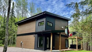 Ein modernes, zweistöckiges Haus mit Holzfassade und schwarzem Dach, umgeben von einem Wald.
