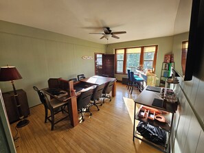 Quarto com mesa de madeira, cadeiras, ventilador de teto e janela com persianas.