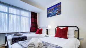 Ein Hotelzimmer mit zwei Betten, einem Fenster mit durchscheinenden Vorhängen, einem Stadtbildgemälde und einem Heizkörper.