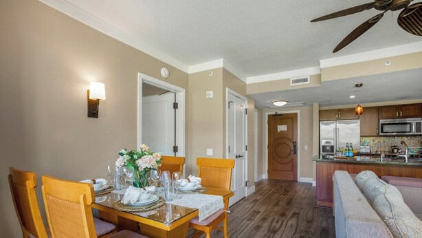 A dining area with a wooden table set for four, a kitchen in the background, and a ceiling fan. - Honua Kai | Ocean View 6 BR, Sleeps 20 | Car Incl. w/ 6+ Nights | HKH ML-2807 by KBM (Lahaina)