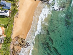 An aerial view of a coastal property with a sandy beach, clear turquoise waters, and a rocky shoreline.