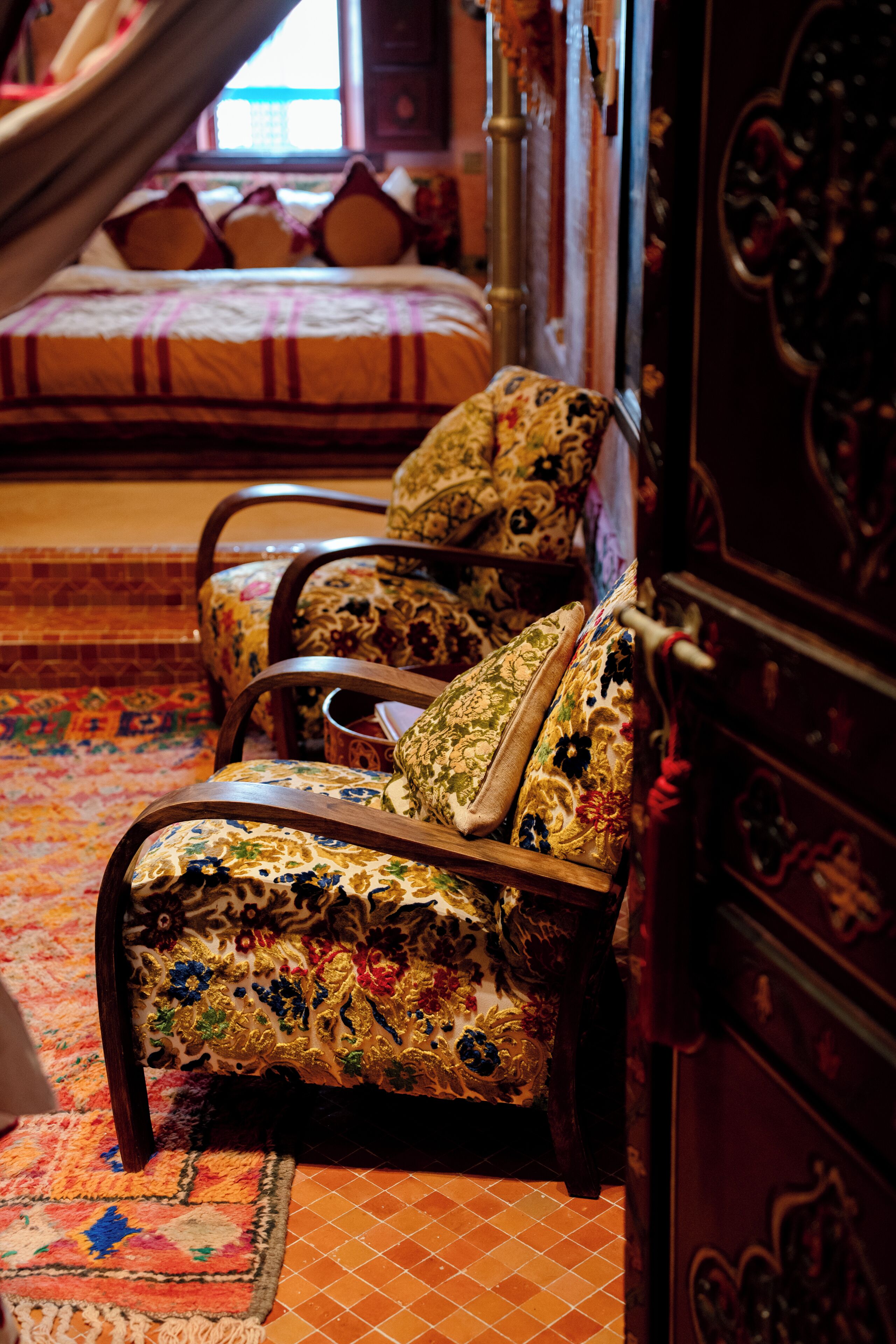 A room with a bed, two ornate chairs, a patterned rug, and a wooden cabinet.