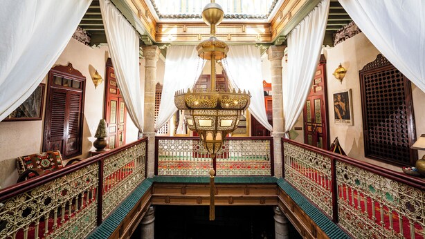 An ornate interior with a central chandelier, a staircase with intricate railings, and traditional decor.