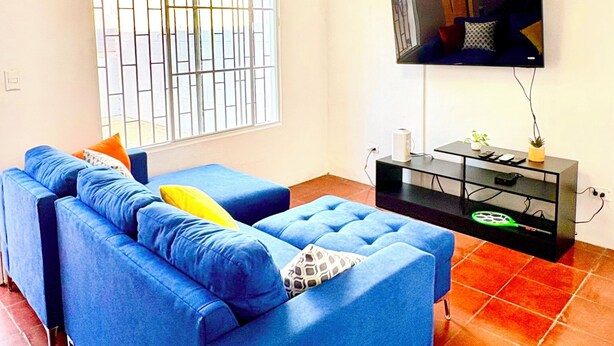 Ein Wohnzimmer mit einem blauen Sofa, einem an der Wand befestigten Fernseher und einem Fenster mit Metallgitter.