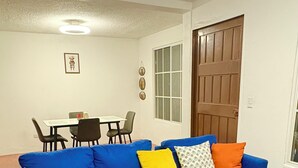 Ein Wohnzimmer mit blauem Sofa, Hocker, Esstisch mit Stühlen, Holztür und Fenster.