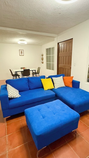 A living room with a blue sofa, an ottoman, a dining table with chairs, a wooden door, and a window. - Fabulous 3-bedroom 2 bath house with AC, gated parking in Santa Ana Department (Santa Ana Department)