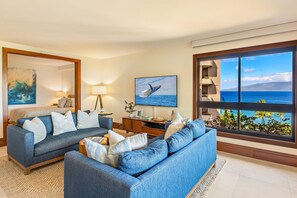 Ein Wohnzimmer mit zwei Sofas, einem Fernseher, einem Gemälde und einem großen Fenster mit Meerblick.