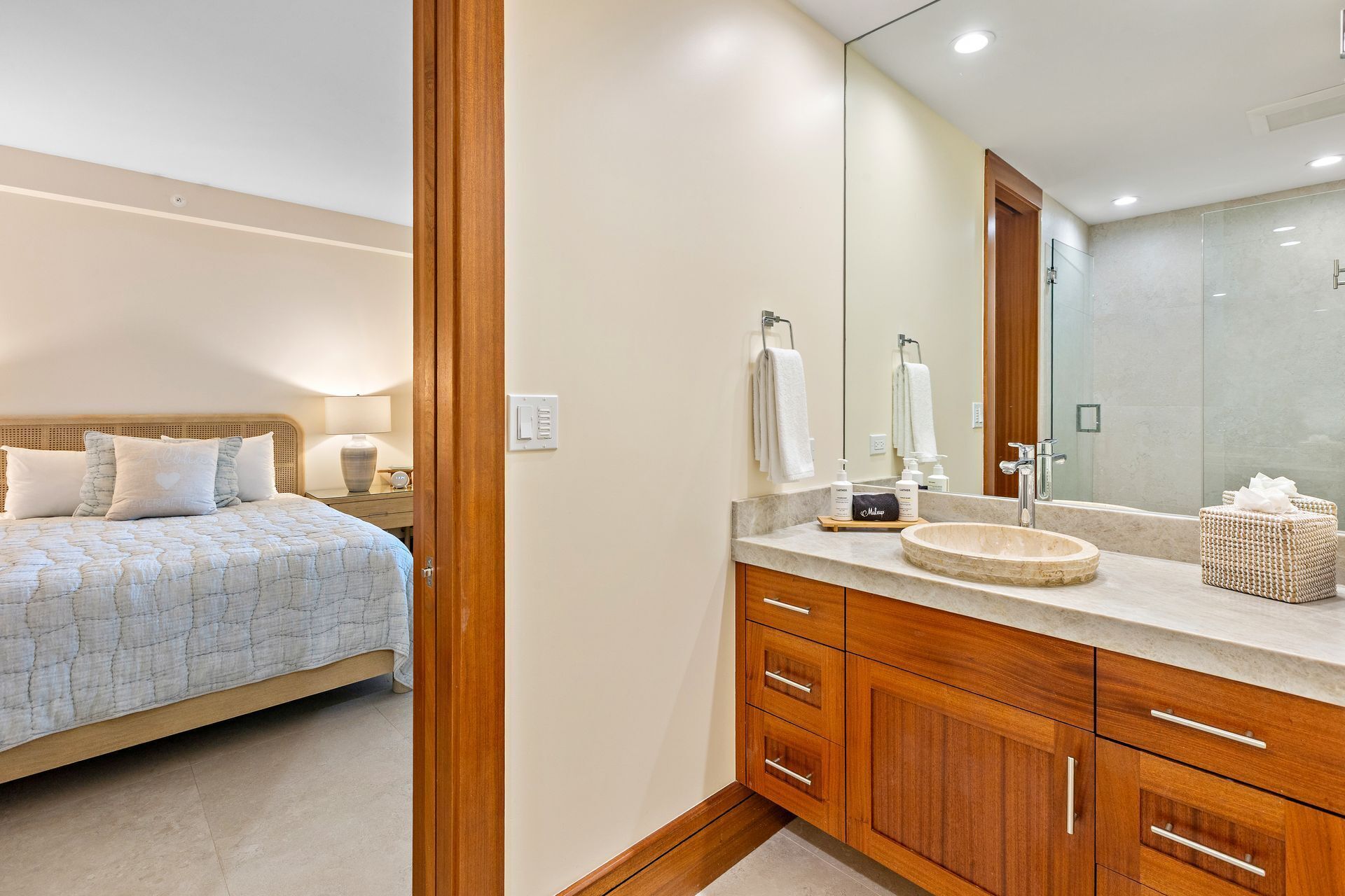 A bedroom with a bed, bedside table, lamp, and a bathroom with a sink, mirror, and towel rack.