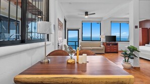 Un espacio habitable amplio con una mesa de comedor de madera, un sofá y un televisor. Amplias ventanas ofrecen vista al océano.