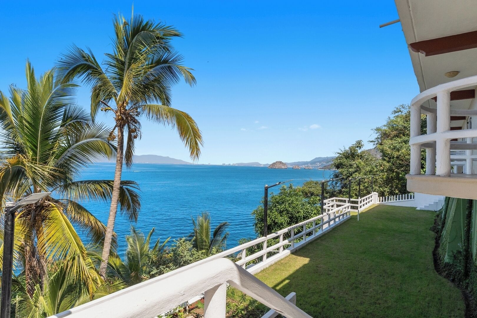 A balcony with a view of the sea, palm trees, and a white fence.