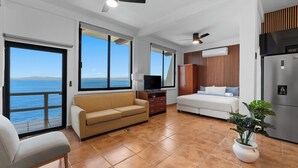 Habitación con sofá, cama, televisión, ventilador de techo, ventana con vista al mar y refrigerador.