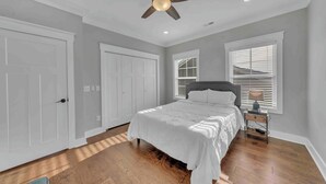 Une chambre à coucher comprenant un lit, une table de chevet, un ventilateur de plafond et deux fenêtres équipées de stores.