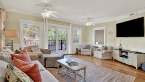 A living room with a sofa, armchairs, a coffee table, and a TV. There are ceiling fans, a window with blinds, and a door leading to an outdoor area. - Charming Tybee Island Escape on the South End | Heated Pool Access (Tybee Island)
