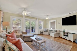 Ein Wohnzimmer mit einer Couch, Sesseln, einem Couchtisch und einem Fernseher. Es gibt Deckenventilatoren, ein Fenster mit Jalousien und eine Tür zu einem Außenbereich.