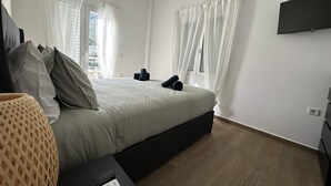 A bedroom with a bed, curtains, a TV, and a window. - Loutra Suites Aegean Wave Suite (Corinth)