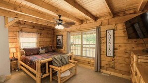 A cabin-style room with a bed, a bench, a ceiling fan, a TV, and a window with curtains. - Mountain Paradise | Mtn View, Hot Tub + FREE Tickets (Sevierville)