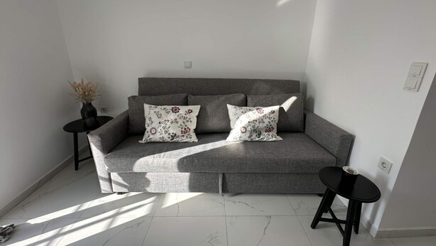 A grey sofa with floral cushions, a small round table with a vase, and a second table with a cup. - Loutra Suites Sunlit Cove Studio (Corinth)