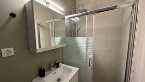 A modern bathroom with a glass shower enclosure, a white sink, and a mirror above it. - Loutra Suites Sunlit Cove Studio (Corinth)