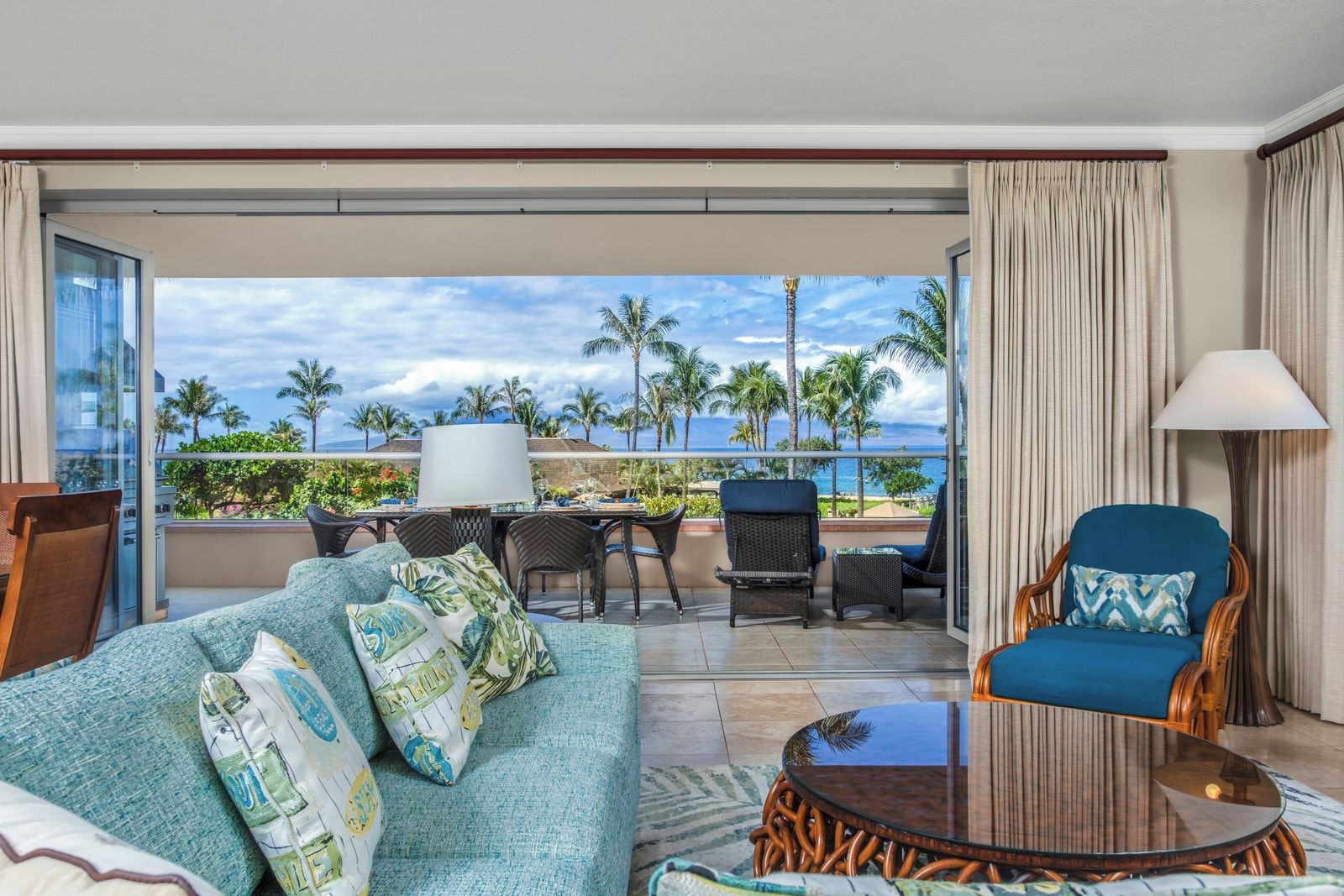 Un balcon aménagé avec un canapé, des fauteuils et une table, donnant sur une plage bordée de palmiers et sur l’océan.