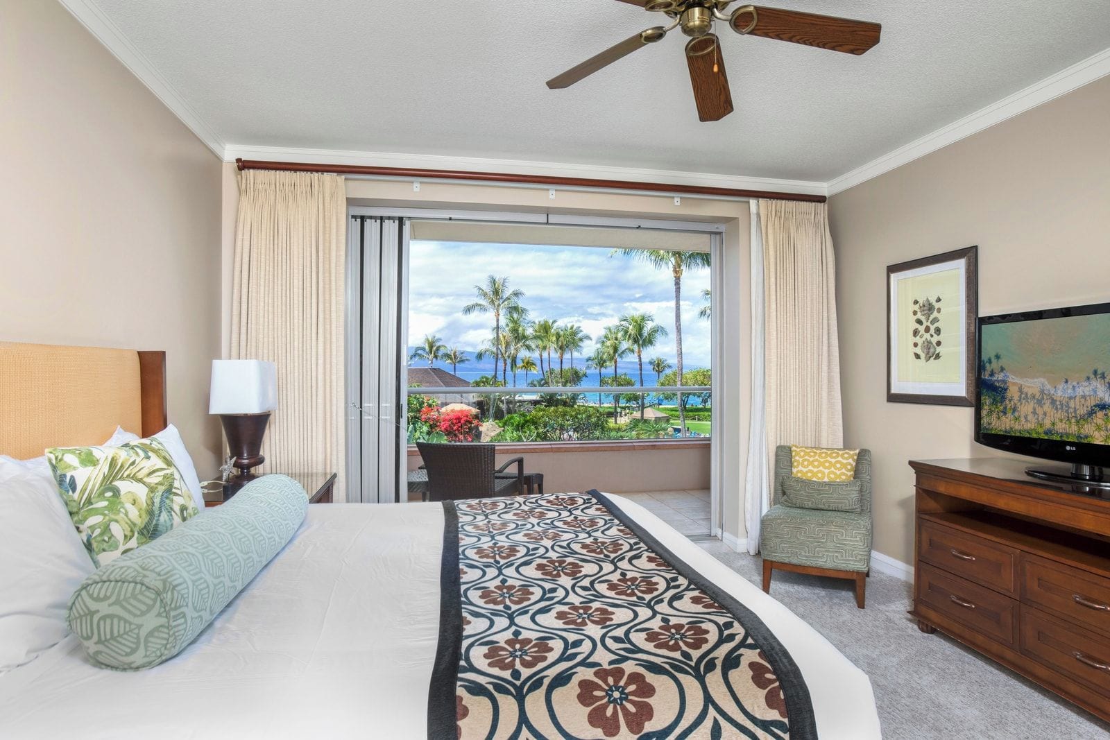 A hotel room with a bed, a television, a ceiling fan, a balcony with a view of palm trees and the ocean, and a chair.
