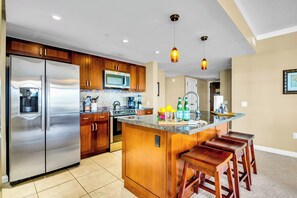 A modern kitchen with stainless steel appliances, wooden cabinets, a central island with bar stools, and pendant lighting. - 3 Honua Kai Villas | Ocean Front | Sleeps 18 | Free Car w/ 6+ Nights | ML-3161 (Lahaina)