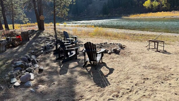 A campsite with Adirondack chairs, a fire pit, and a river view.