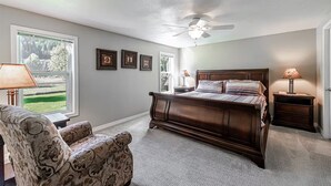 A bedroom with a large bed, a chair, a nightstand, a ceiling fan, and framed pictures on the wall.