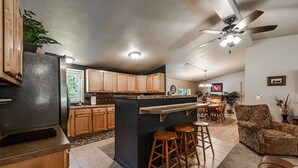 A modern kitchen with wooden cabinets, a central island with stools, a ceiling fan, and a dining area with a patterned sofa.
